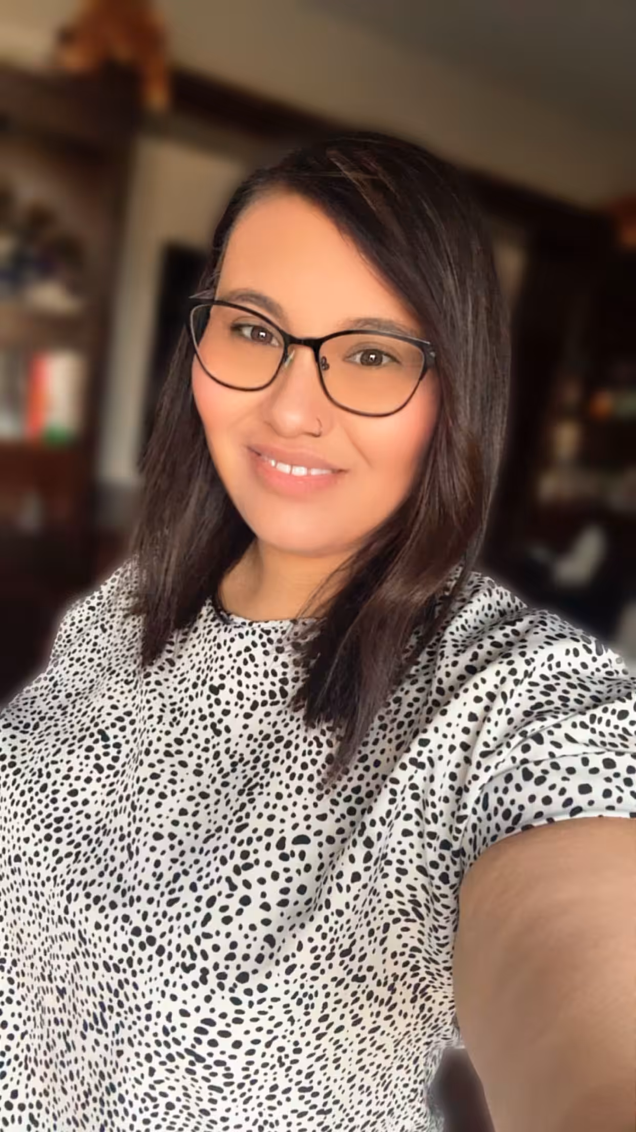 A woman with straight, dark hair and glasses smiles at the camera. She is wearing a white shirt with black polka dots. The background shows a blurred room with shelves and books.