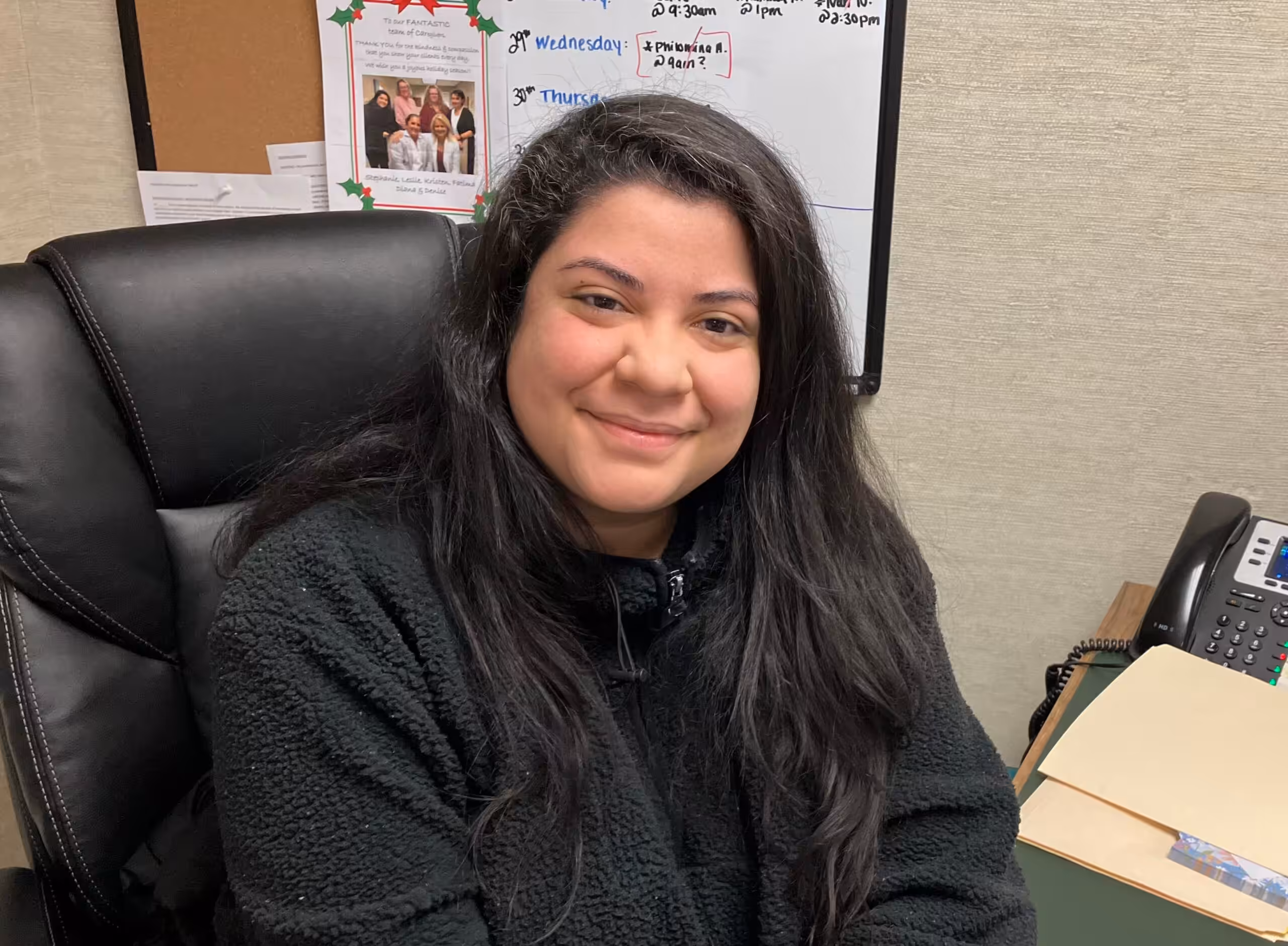 A woman with long dark hair and a black fleece jacket sits and smiles at a desk. Behind her is a whiteboard with notes, a photo, and some paperwork, as well as a phone and folders on the desk.