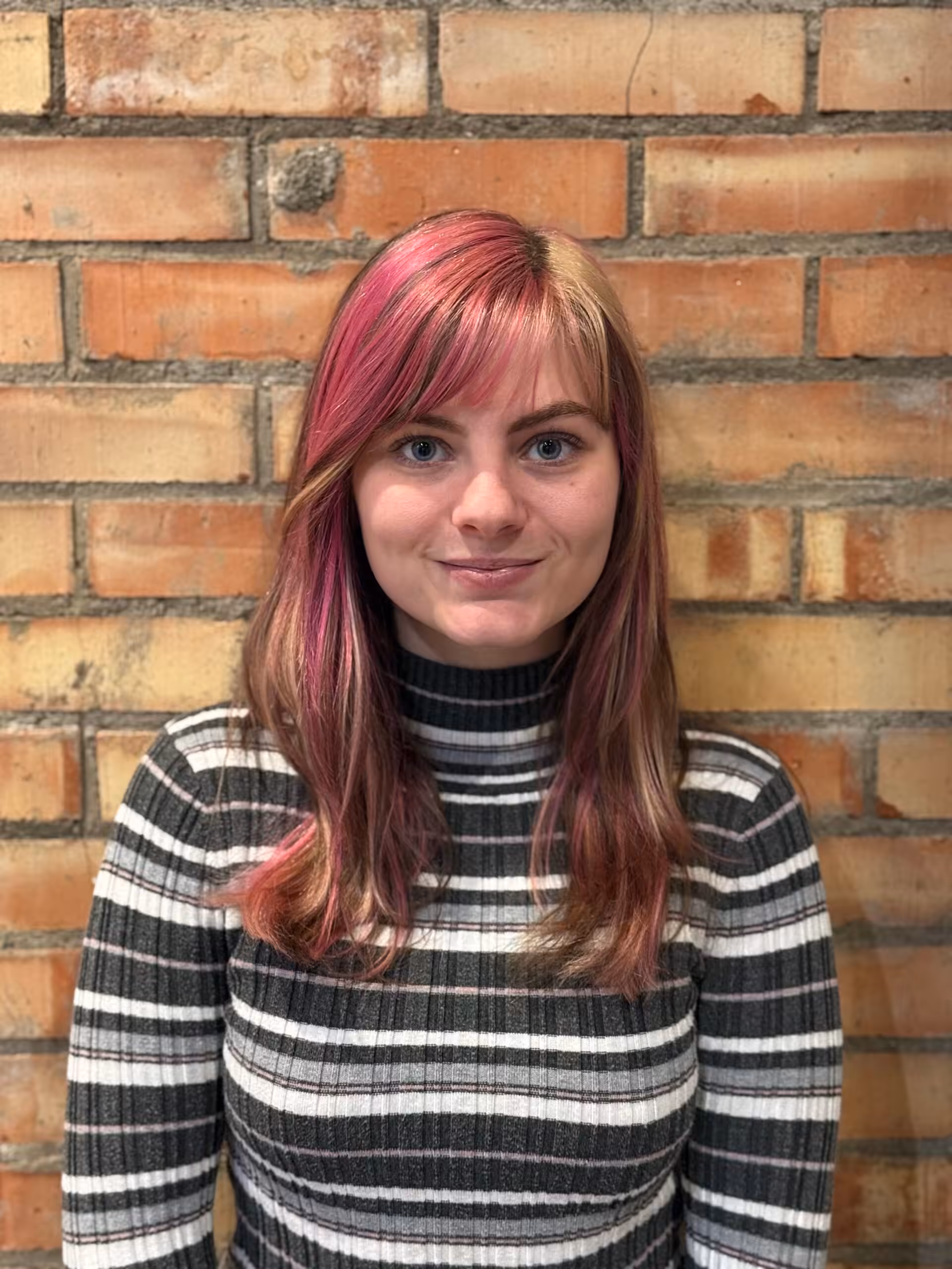 A person with straight, shoulder-length brown hair with pink highlights stands in front of a brick wall, wearing a black and white striped turtleneck sweater and smiling slightly.