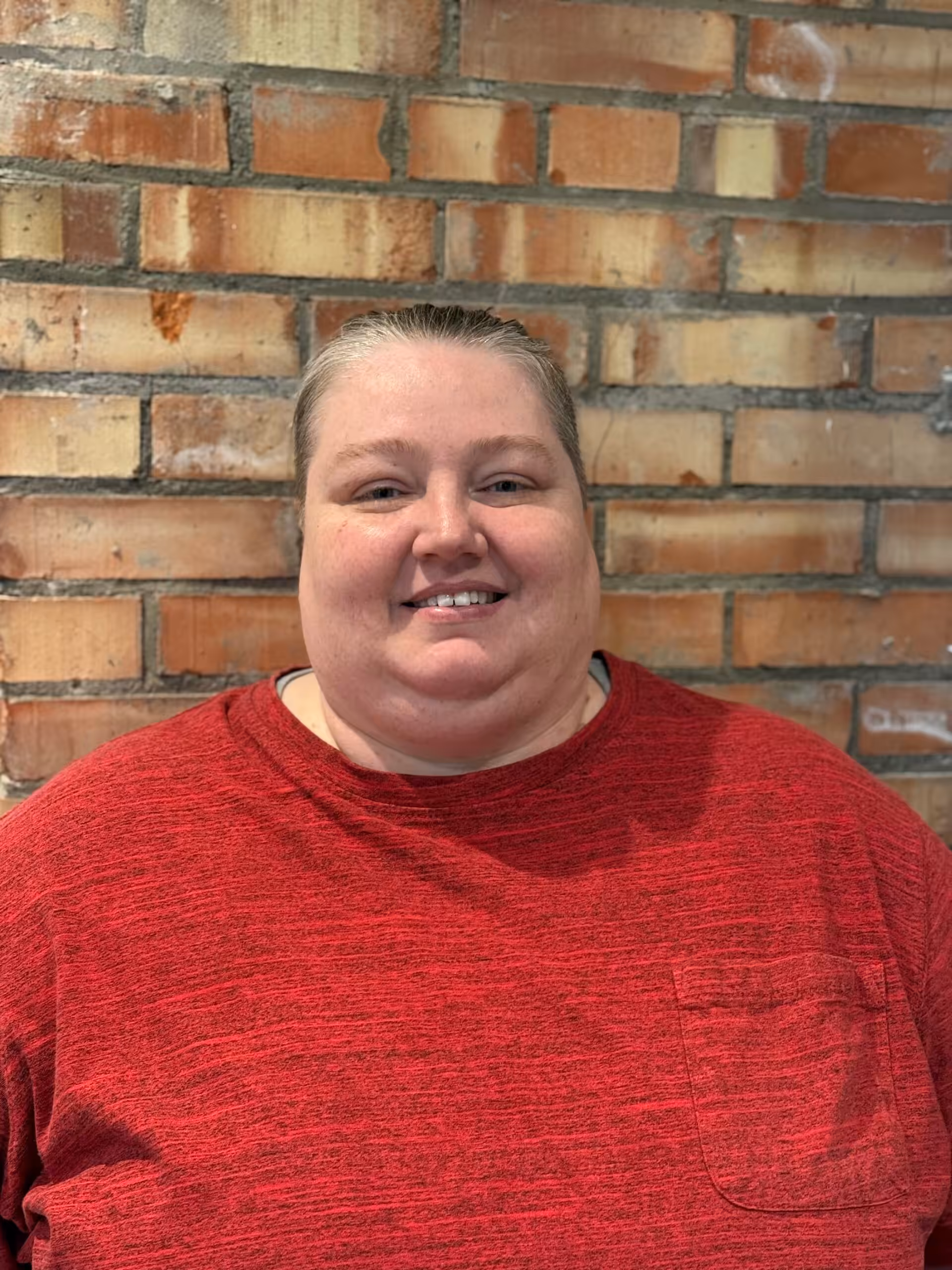 A person with short, light hair smiles at the camera while standing in front of a brick wall. They are wearing a red shirt with a pocket on the left side.