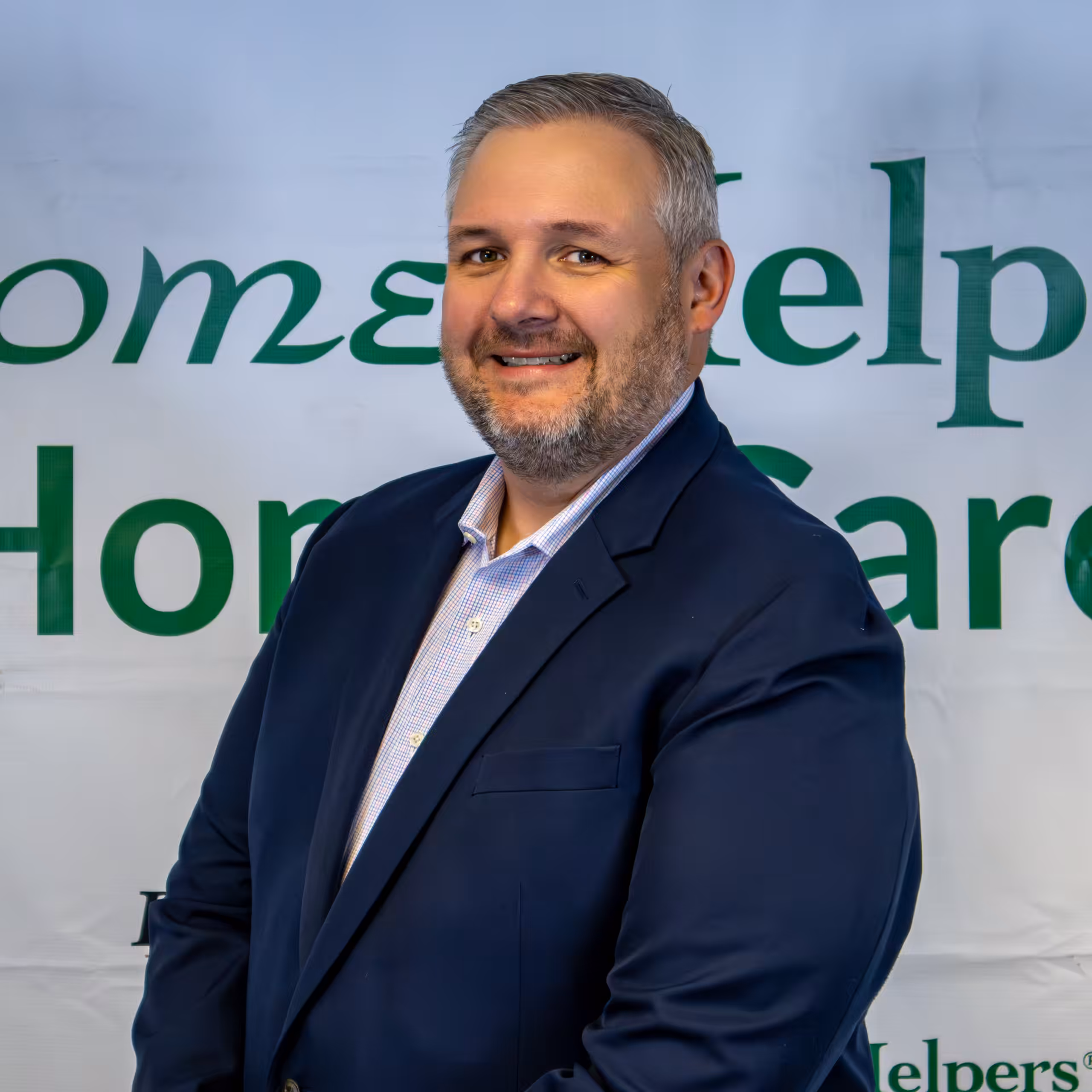 A man with short gray hair and a trimmed beard, wearing a navy suit and light shirt, stands smiling in front of a banner that says Home Helpers Home Care.