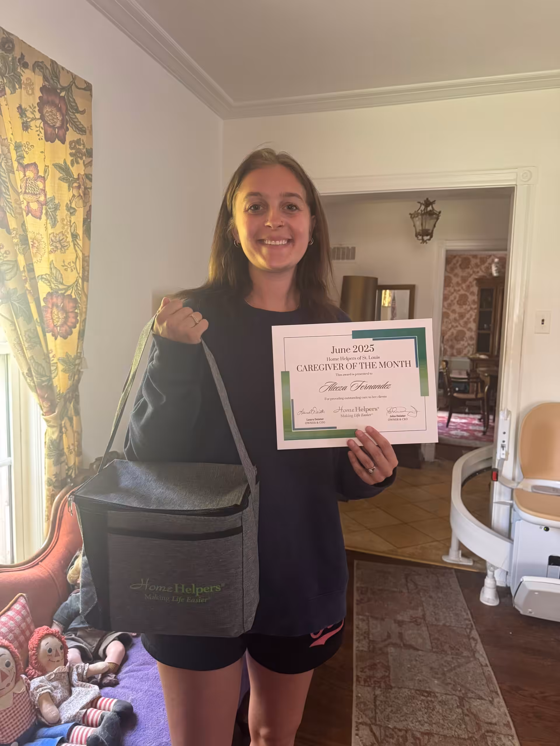 A smiling woman stands indoors holding a “Caregiver of the Month” certificate for June 2025 in one hand and a gray Home Helpers bag in the other. There are dolls on a chair and floral curtains in the background.