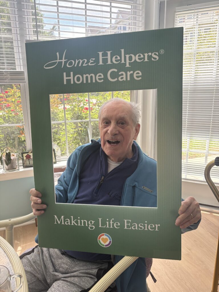 An elderly man smiles while holding a large photo frame sign that reads Home Helpers Home Care - Making Life Easier in a sunlit room with windows and plants in the background.