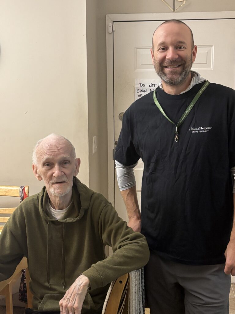 Two men indoors: one seated older man in a green hoodie, and a standing man smiling, wearing a black shirt and lanyard. A white door with a note is in the background.