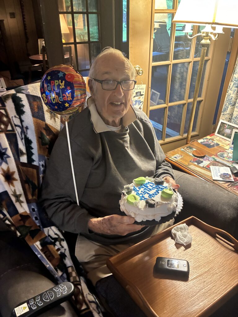 An elderly man smiles while holding a birthday cake and a Happy Birthday balloon, sitting in a cozy living room with a patterned blanket on his chair and a remote control on a tray table nearby.
