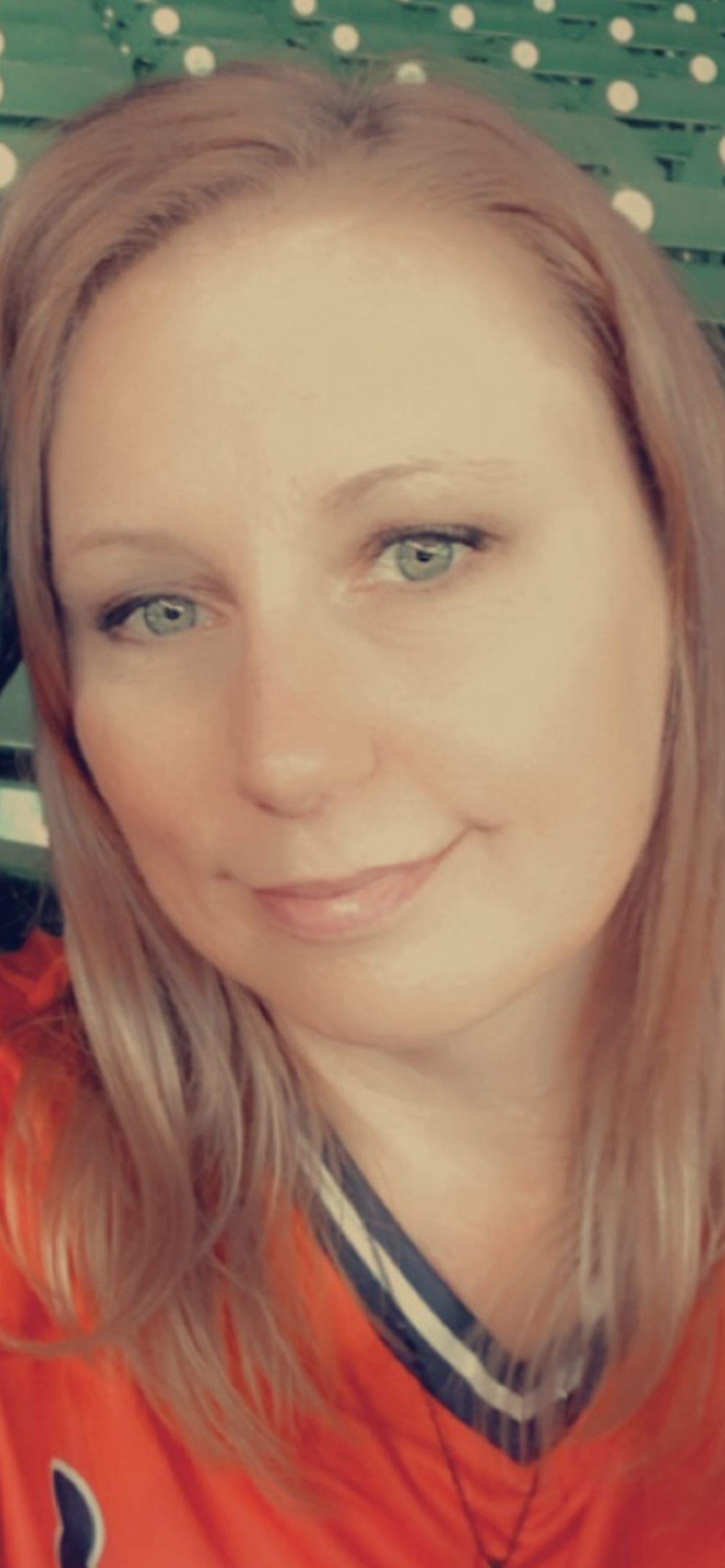 A woman with straight, light brown hair and green eyes smiles softly at the camera. She is wearing an orange top and is seated in front of green bleachers with white dots.