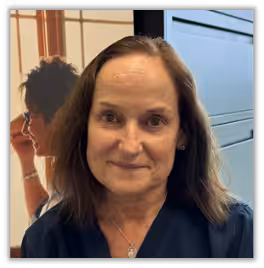A woman with shoulder-length brown hair and a navy top smiles softly at the camera. In the background, there is a blurred image of another person and a filing cabinet.