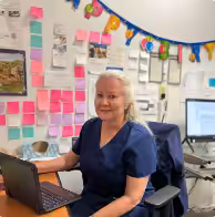 A woman with long gray hair, wearing navy blue scrubs, sits at a desk with a laptop. Behind her, colorful sticky notes and a “Happy Birthday” banner decorate the wall.