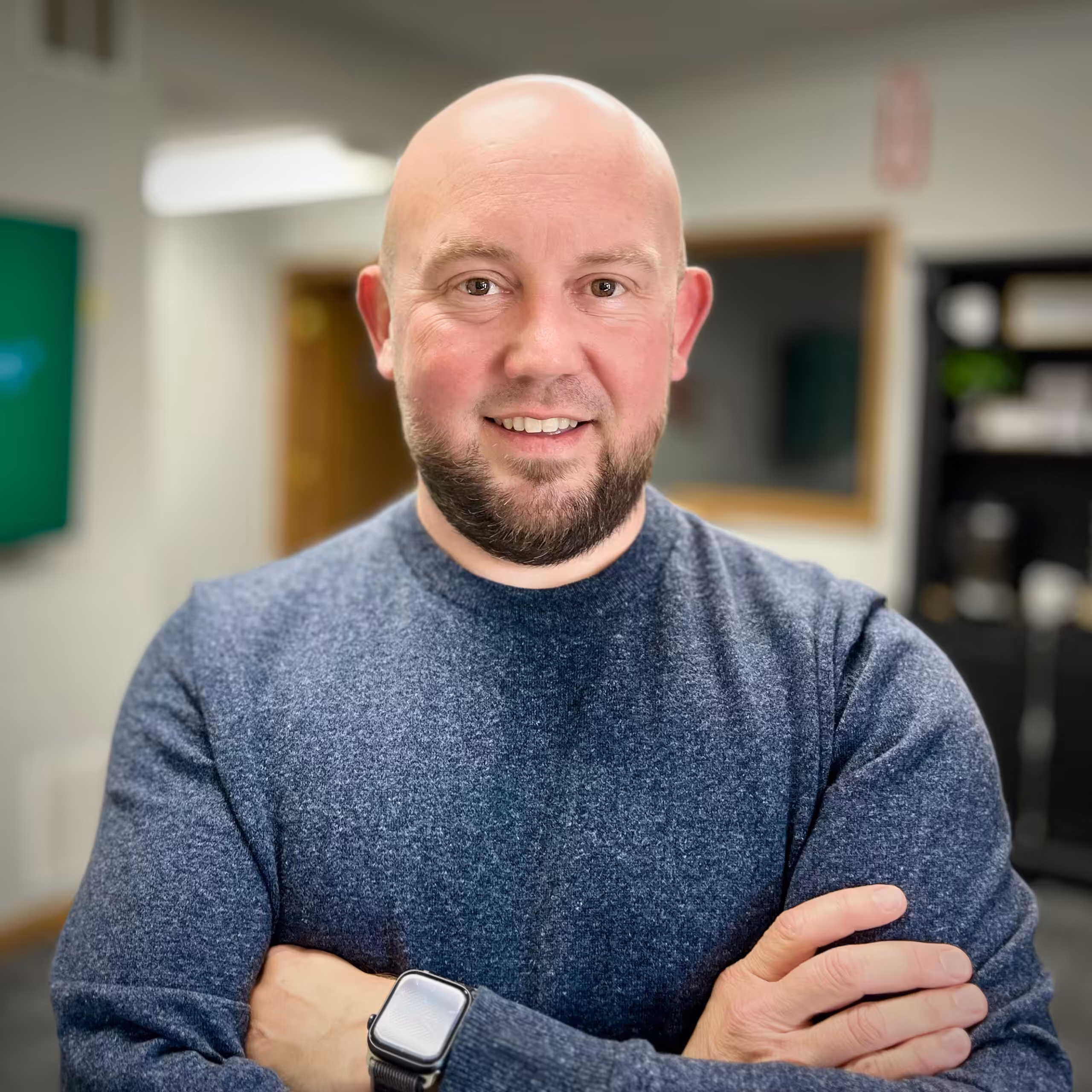 A bald man with a beard, wearing a gray sweater and a smartwatch, stands indoors with his arms crossed, smiling at the camera. Shelves and a doorway are visible in the blurred background.