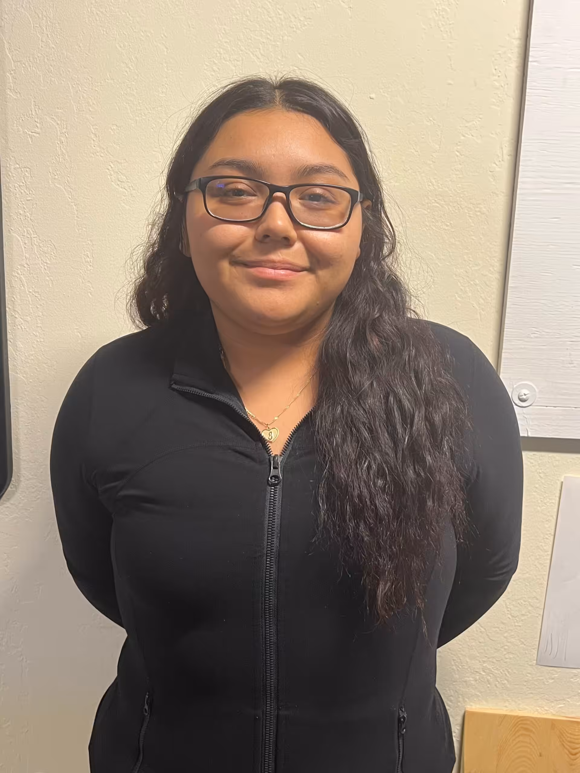 A woman with long wavy dark hair and glasses stands in front of a light-colored wall, wearing a black zip-up top and smiling slightly at the camera.