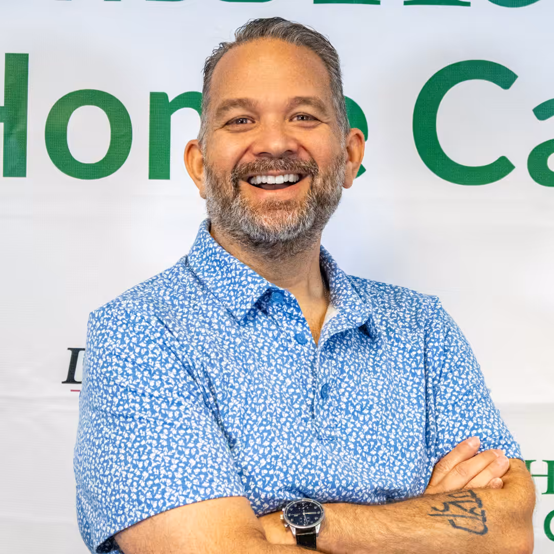 A smiling man with a short beard and mustache wearing a blue patterned shirt stands with arms crossed in front of a white banner with green text. He has a watch on his wrist and a visible tattoo on his forearm.