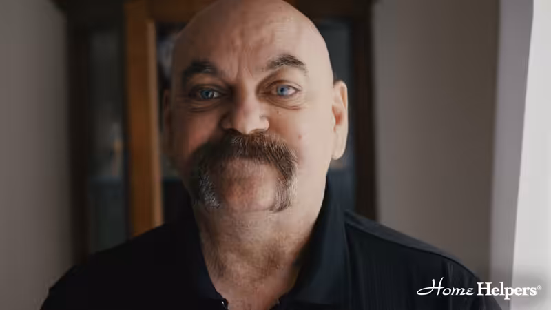 A bald man with a thick, dark mustache and blue eyes smiles slightly, wearing a black shirt. The background shows a wooden cabinet. The Home Helpers logo is in the bottom right corner.