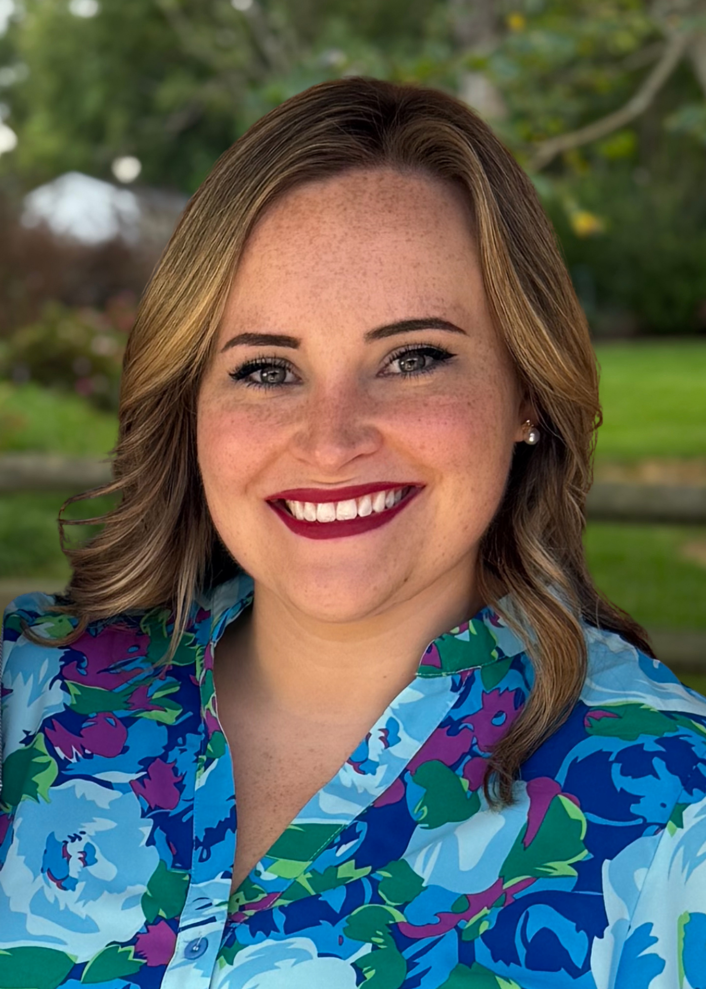 A woman with wavy light brown hair smiles outdoors, wearing a blue floral blouse. The background features green grass, trees, and a wooden fence, softly blurred.
