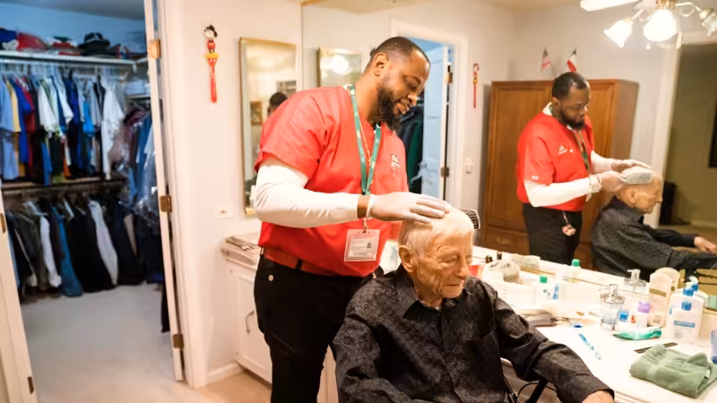 A caregiver in red scrubs gently helps an elderly man seated in front of a bathroom mirror. The room is well-lit, with toiletries on the counter and an open closet visible in the background.