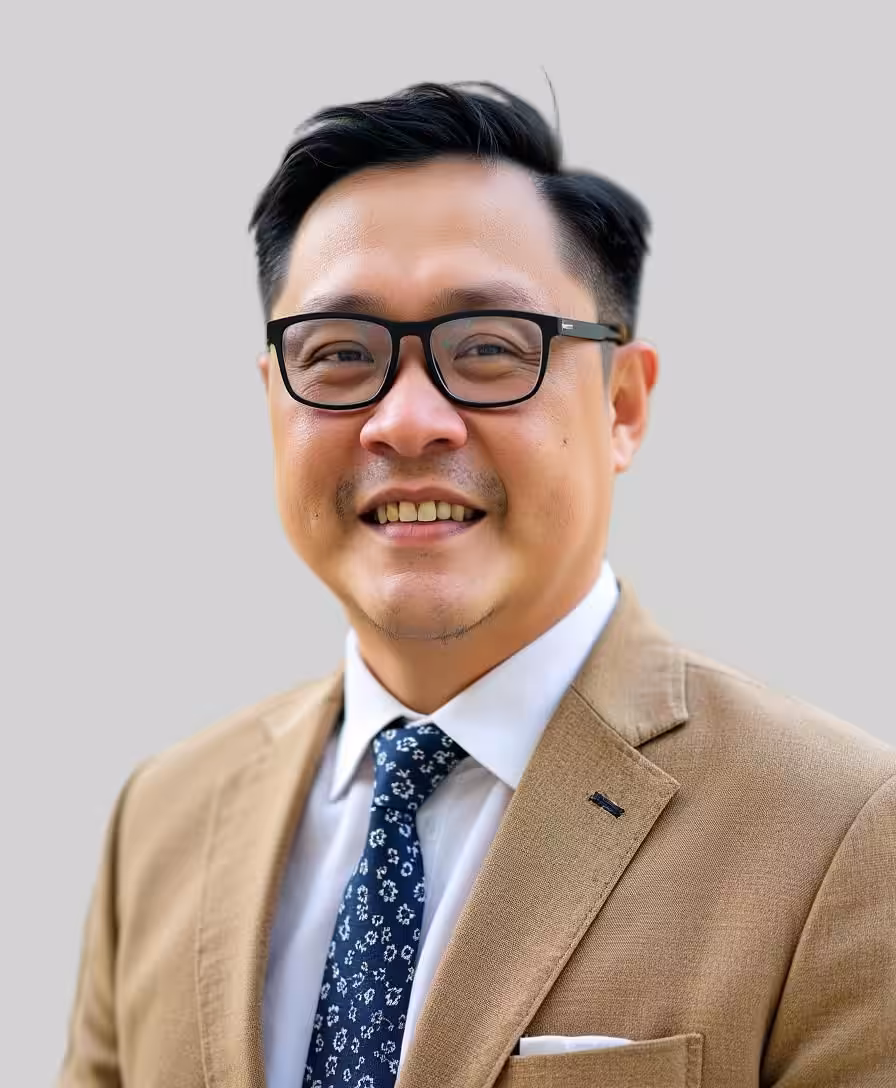 A man with short black hair and glasses is smiling. He is wearing a tan suit jacket, a white shirt, and a patterned navy tie. The background is plain light gray.