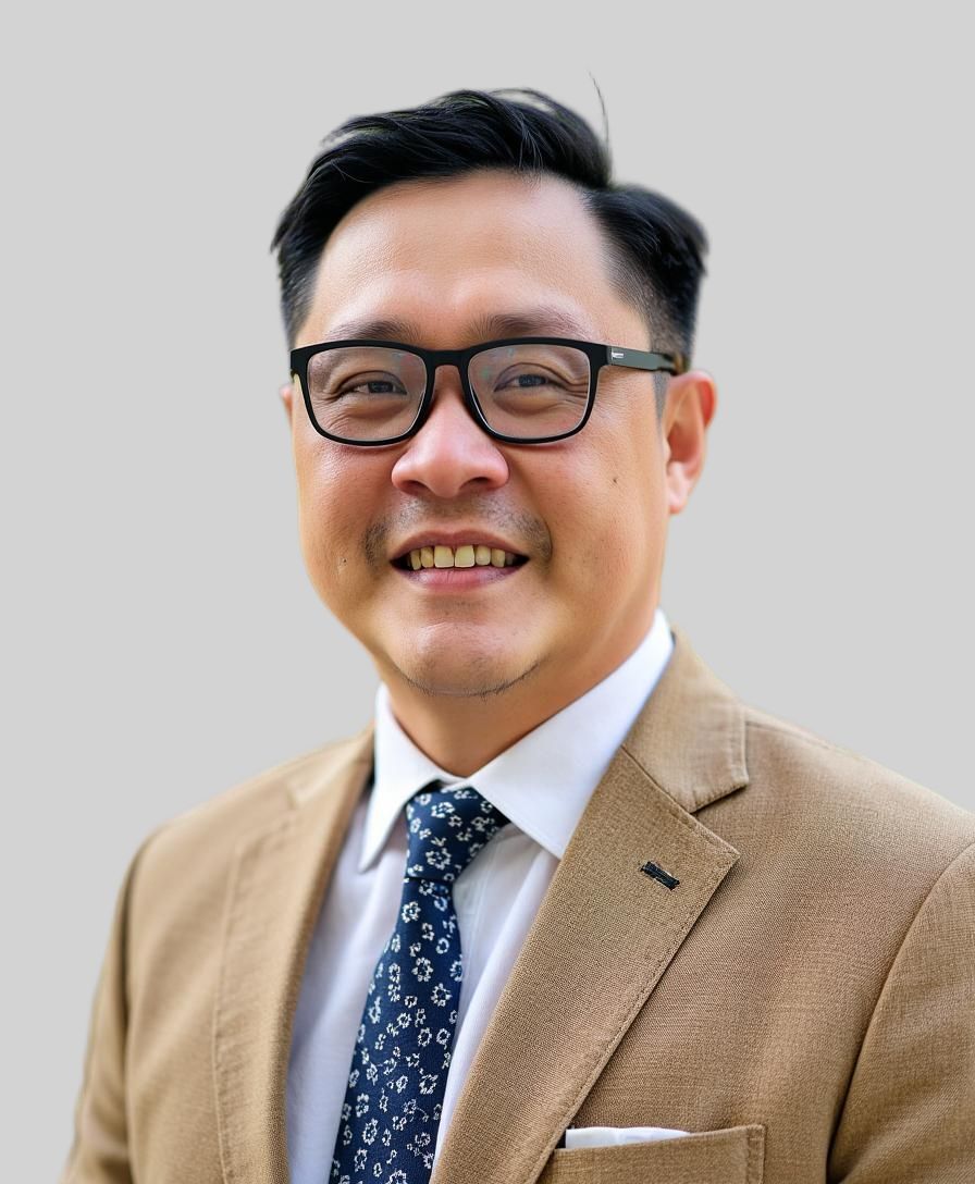 A man with short black hair and glasses is smiling. He is wearing a tan suit jacket, a white shirt, and a patterned navy tie. The background is plain light gray.