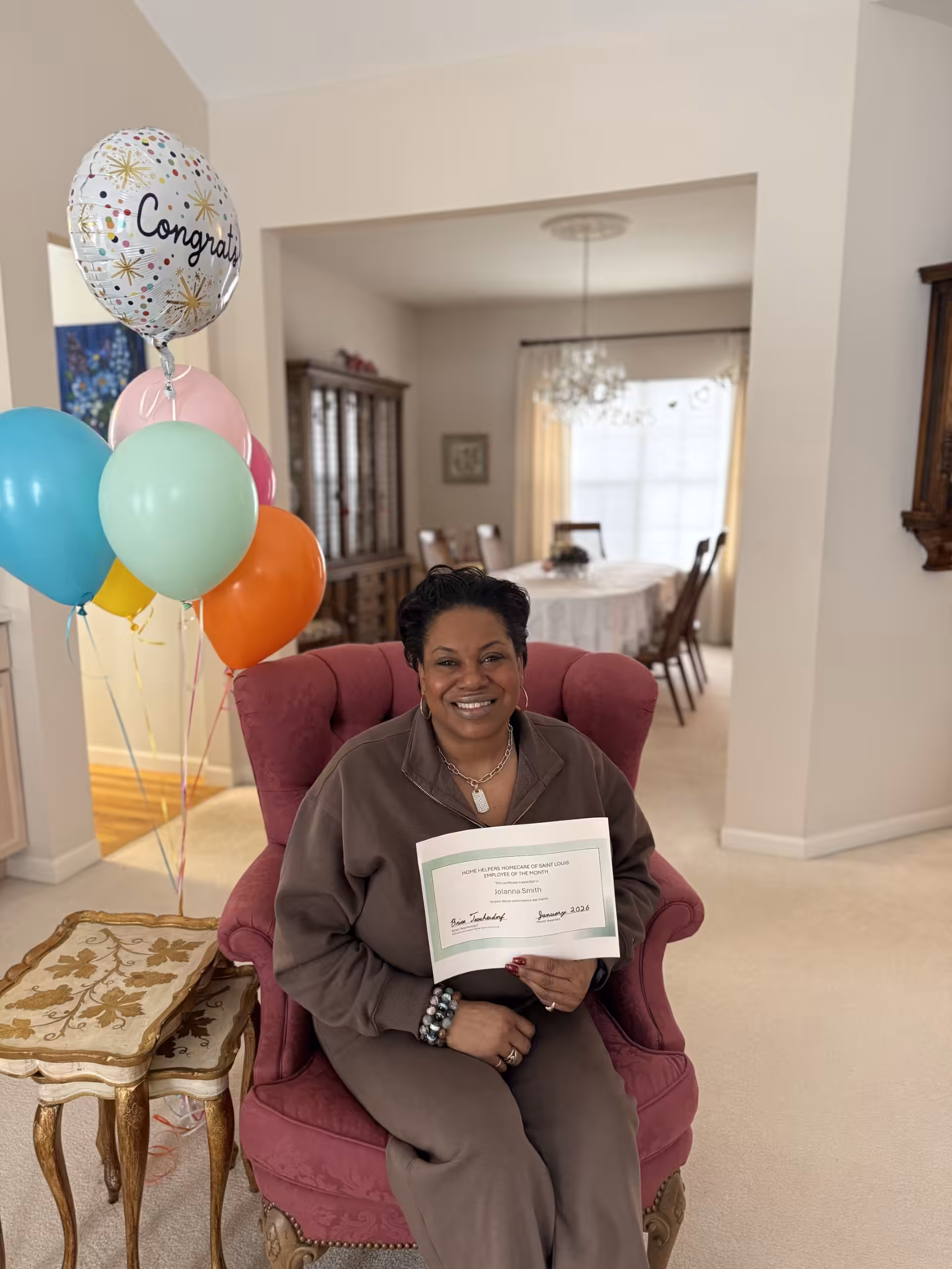 A woman sitting in a pink chair, smiling and holding a certificate. Colorful balloons, including one that says Congrats, are beside her. The setting is a bright, neatly decorated living room with a dining area in the background.
