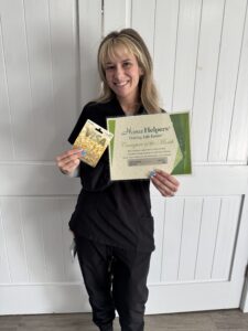 A woman in black scrubs smiles while holding a gift card and a “Caregiver of the Month” certificate from Home Helpers in front of white paneled doors.
