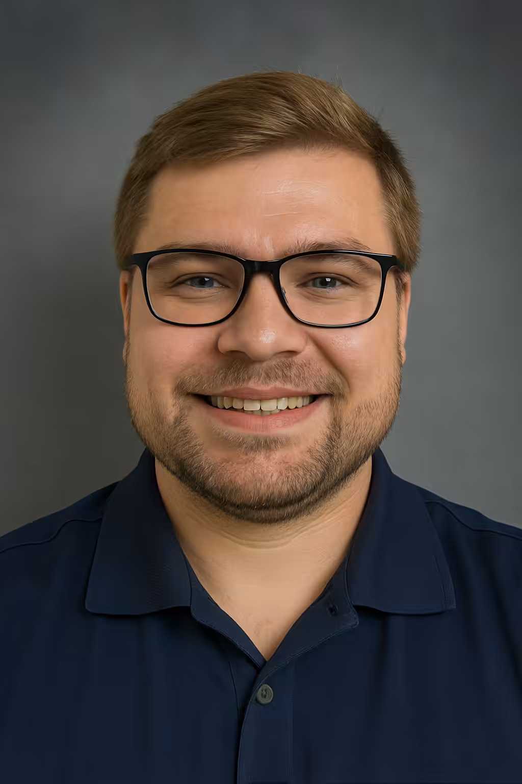 A smiling man with light brown hair, a short beard, and black glasses, wearing a navy blue collared shirt, poses in front of a plain gray background.