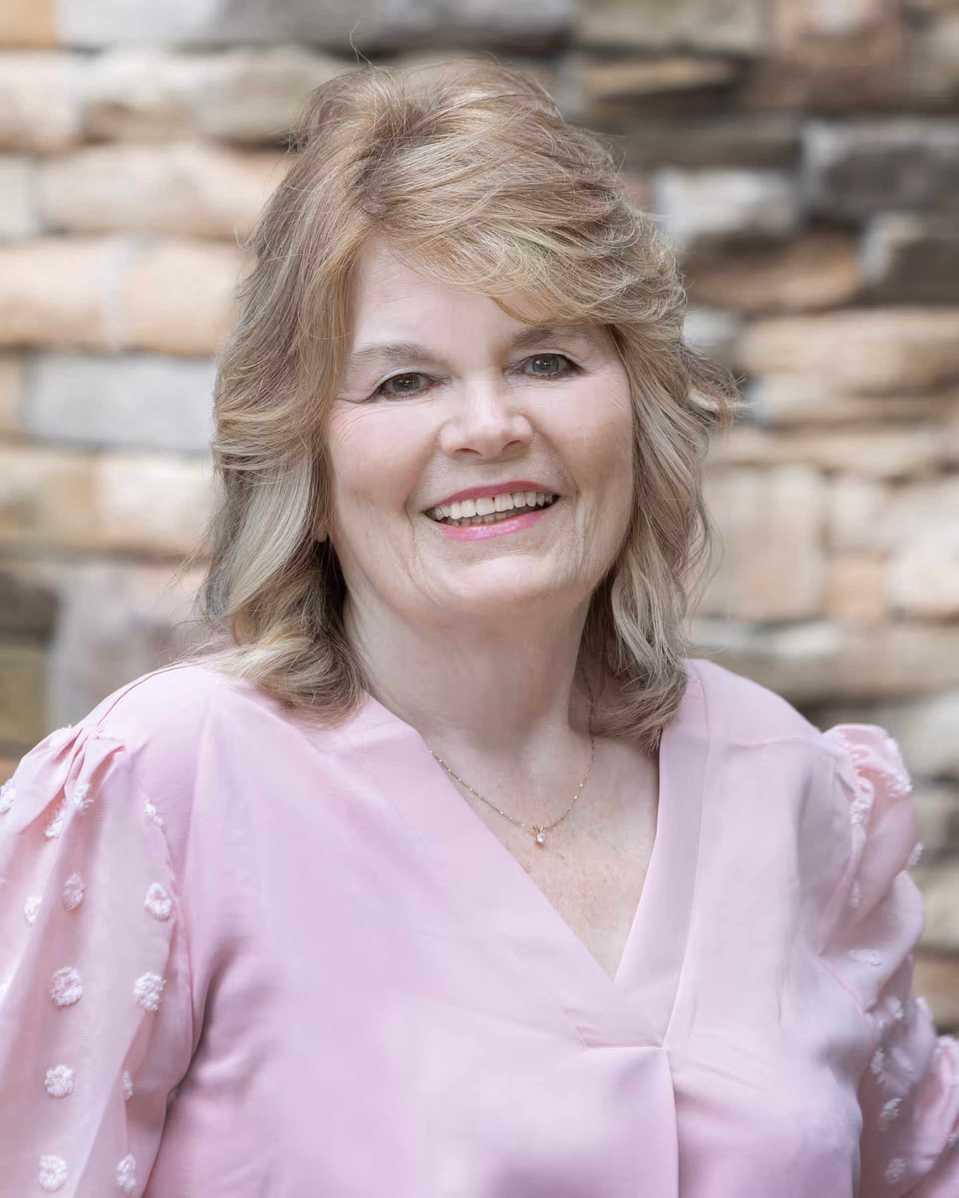 A smiling older woman with shoulder-length blonde hair stands in front of a stone wall, wearing a light pink blouse with sheer sleeves.