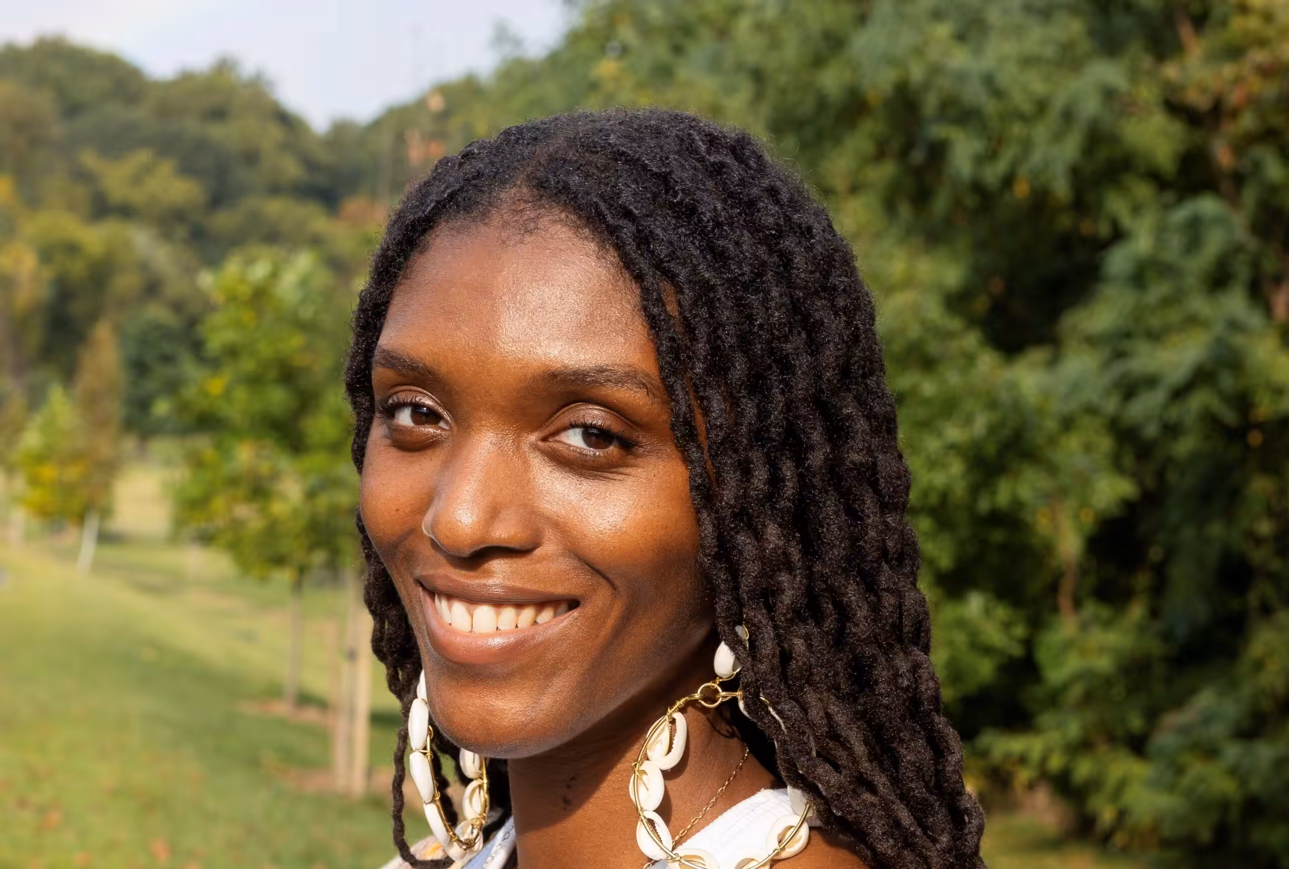 A woman with long, twisted hair and shell earrings smiles outdoors in a green park with trees in the background.