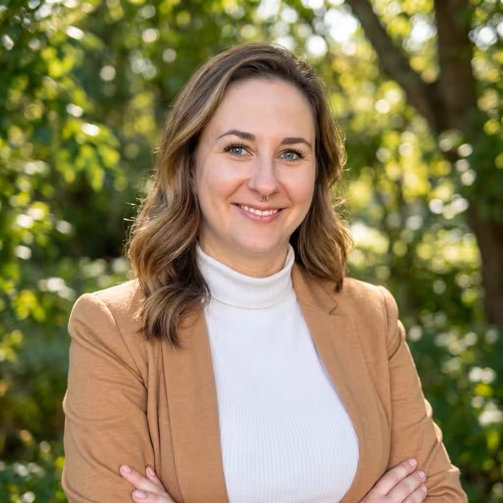 A woman with wavy brown hair and a nose ring is smiling, standing outdoors with her arms crossed. She is wearing a tan blazer over a white turtleneck. Green trees and sunlight are visible in the background.