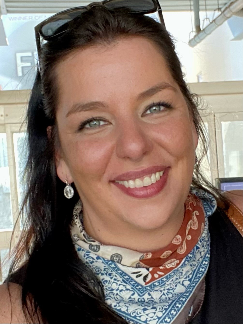 A close-up portrait of a smiling woman wearing a patterned neck scarf and sunglasses on her head.
