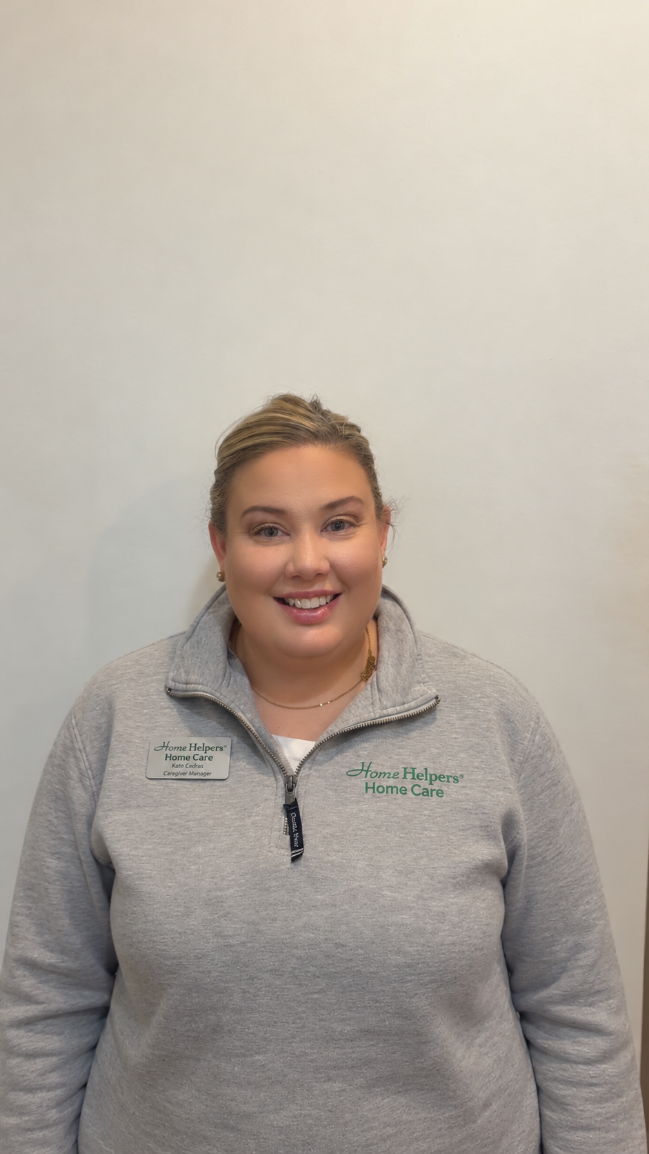 A smiling woman wearing a grey Home Helpers Home Care zip-up sweater and a name tag.