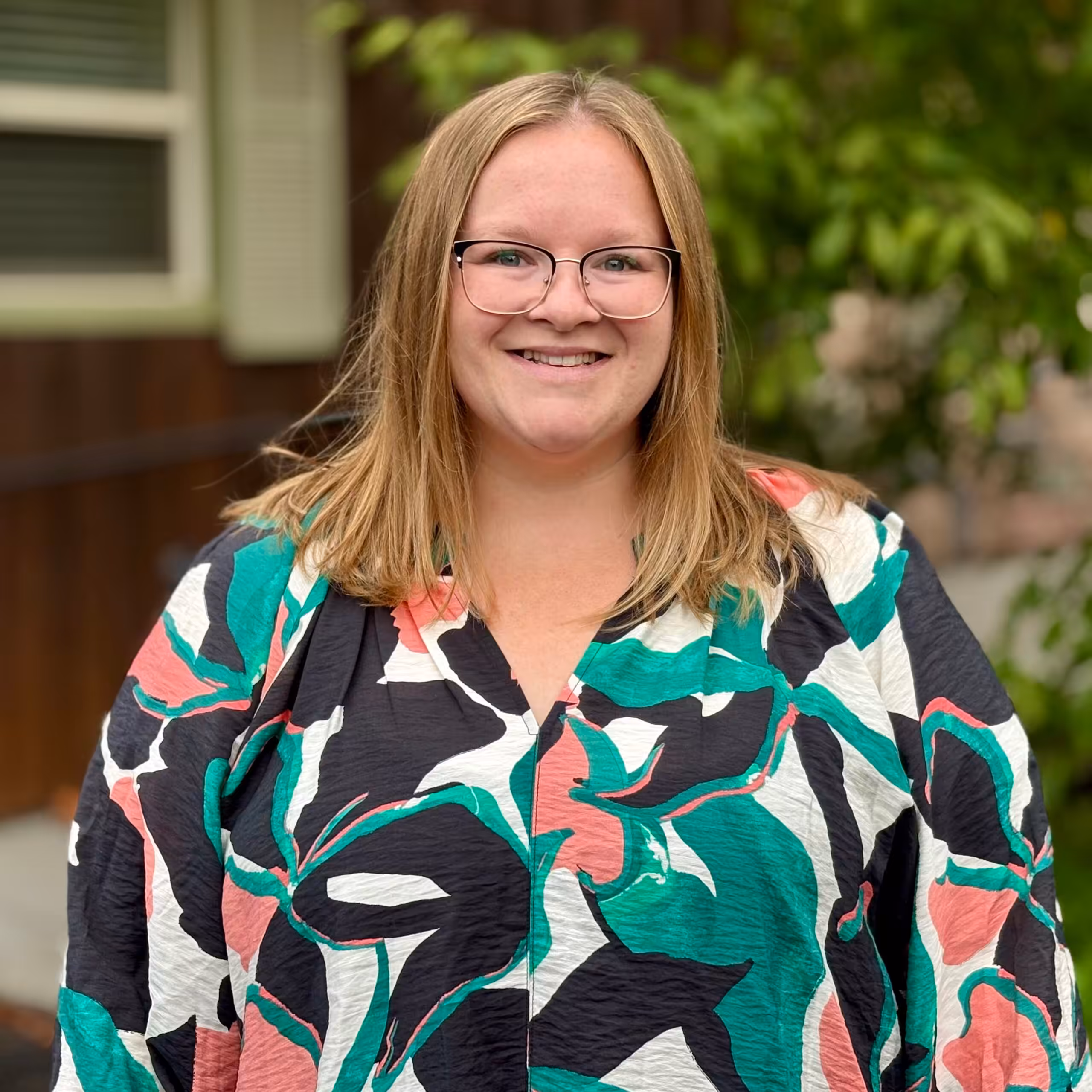 A smiling woman with straight, shoulder-length blonde hair and glasses wears a colorful, abstract-patterned top. She stands outside in front of greenery and a building with a window and brown siding.