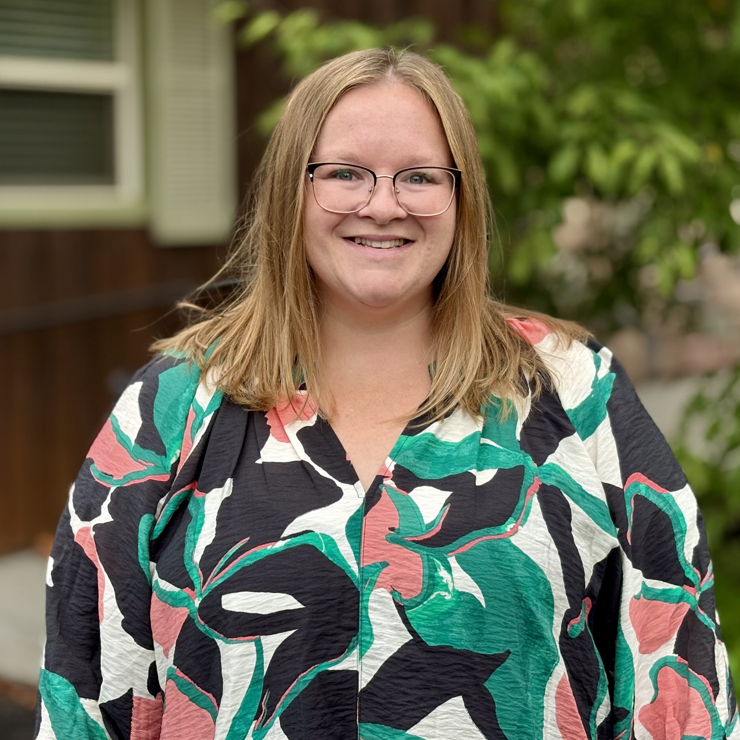 A smiling woman with straight, shoulder-length blonde hair and glasses wears a colorful, abstract-patterned top. She stands outside in front of greenery and a building with a window and brown siding.