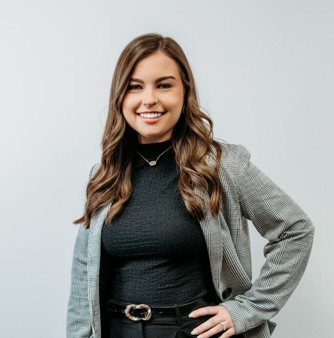 A woman with long brown hair, wearing a black top, plaid blazer, and black pants, stands smiling against a plain light background. One hand rests on her hip.