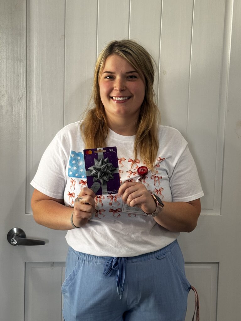 A smiling woman with blonde hair, wearing a white shirt with orange designs and blue pants, stands in front of a white door holding two gift cards, one with a silver bow and one blue with white stars.