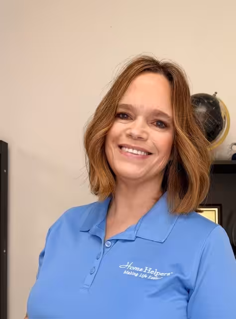 A woman with shoulder-length brown hair smiles while wearing a blue Home Helpers polo shirt; a black shelf and globe are visible in the background.