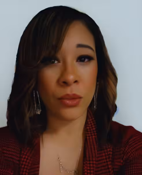 A woman with shoulder-length brown hair, wearing a red patterned top, dangling earrings, and a necklace, looks directly at the camera against a plain light background.