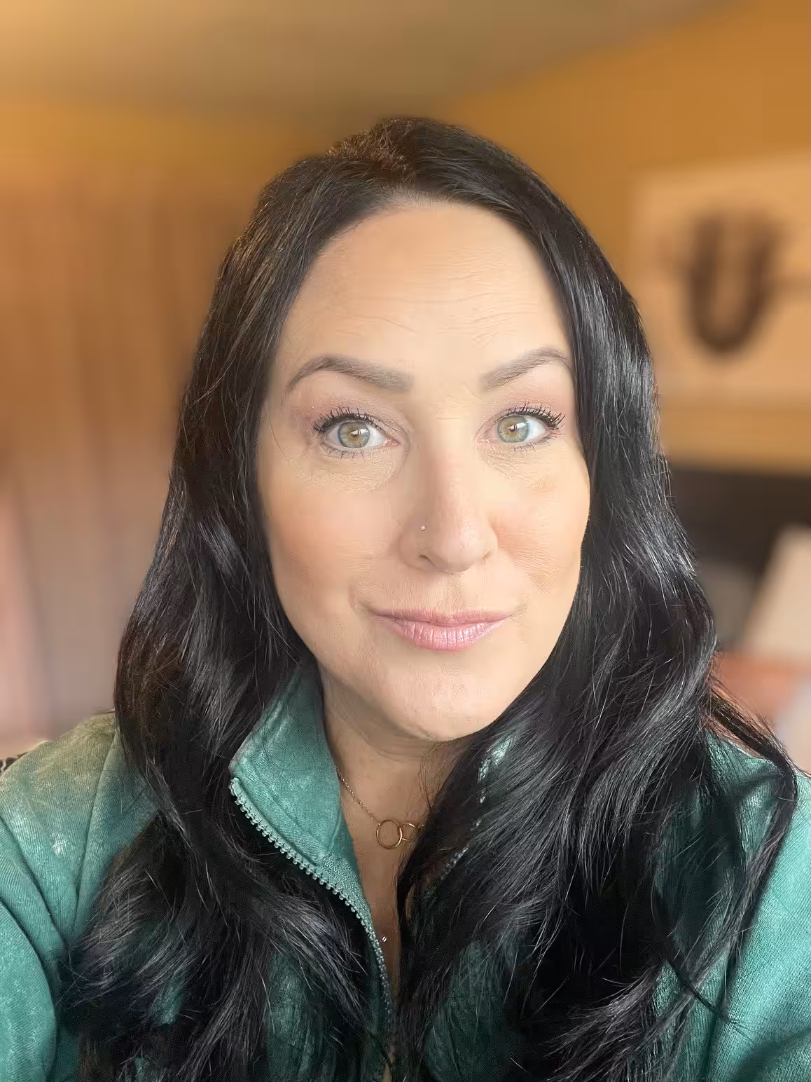 A woman with long black hair and green eyes smiles softly at the camera. She is wearing a green zip-up top and a delicate necklace. The background appears to be a warmly lit room with blurred decor.