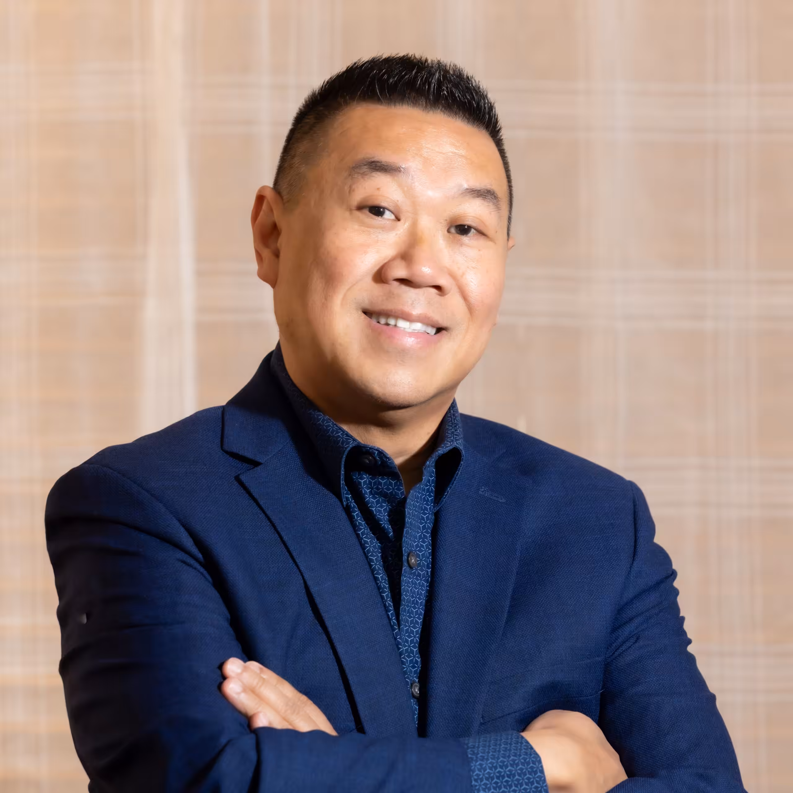 A man wearing a dark blue suit and patterned shirt stands with his arms crossed, smiling confidently in front of a light-colored, plaid background.