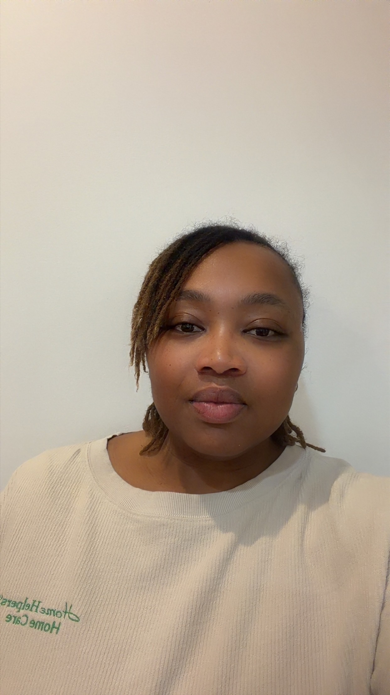 A person with locs wearing a cream-colored shirt with green embroidered text that reads Home Helpers Home Care.