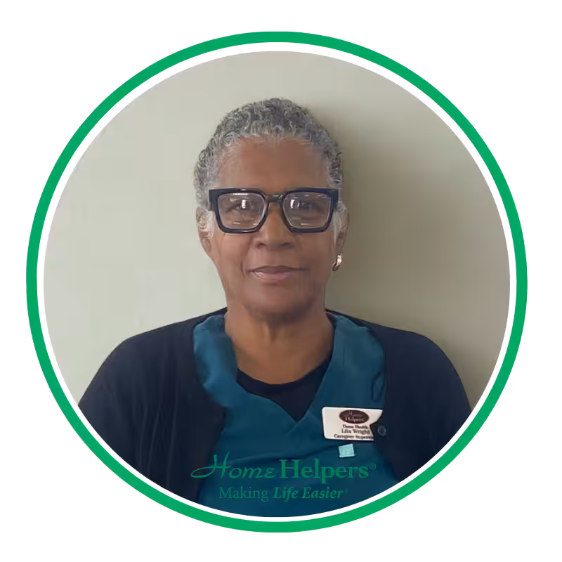 A woman with short gray hair and glasses, wearing a dark sweater and teal Home Helpers uniform, sits against a light wall and smiles slightly. She has a name badge on her left side.