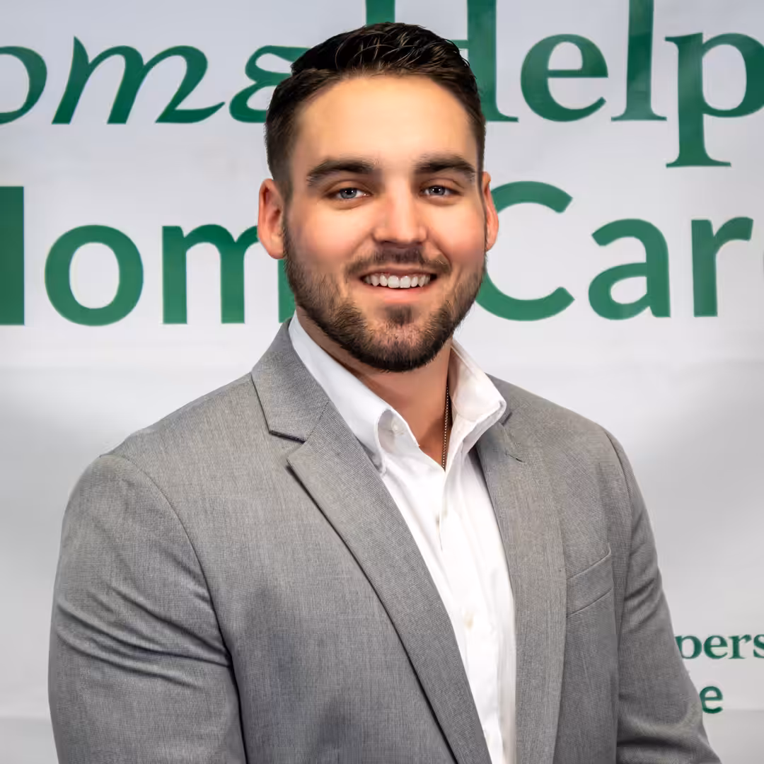 A man with short dark hair and a trimmed beard, wearing a light gray blazer and white shirt, stands smiling in front of a banner with green text that reads “Help Home Care.”.