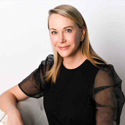 A woman with straight blonde hair, wearing a black top with sheer sleeves, sits on a light-colored chair and smiles at the camera against a white background.
