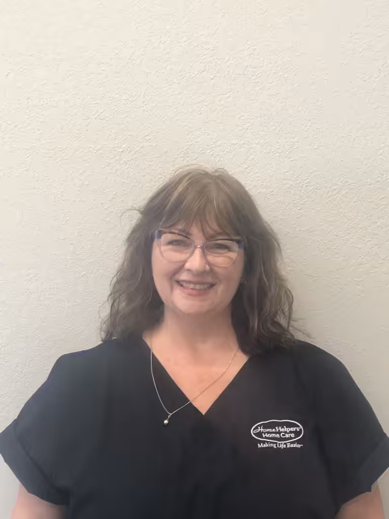 A woman with wavy brown hair and glasses smiles at the camera. She is wearing a black scrub top with an embroidered logo that reads Home Helpers Home Care, Making Life Easier and a necklace. The background is a plain white wall.
