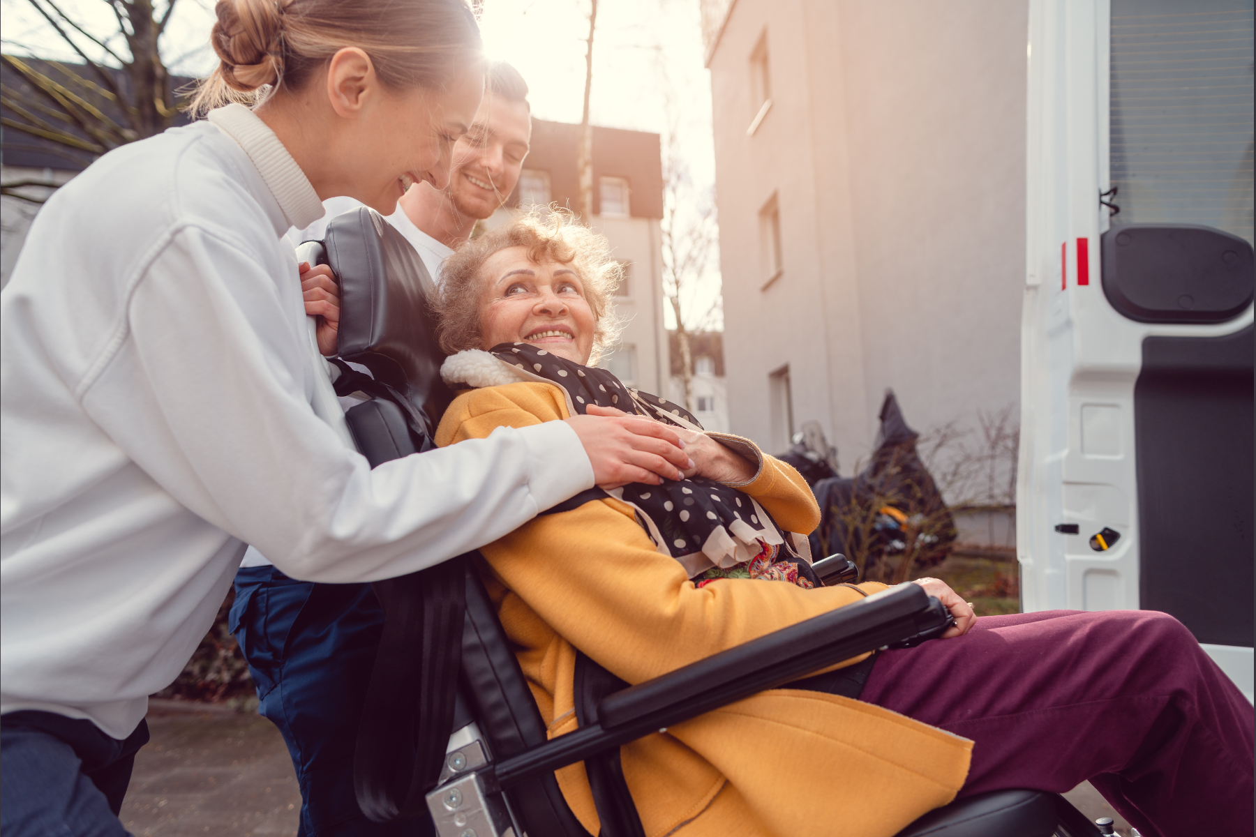 A smiling elderly woman in a wheelchair is being assisted by a caregiver and a man outdoors near a vehicle. The group looks happy and engaged, with sunlight shining in the background.