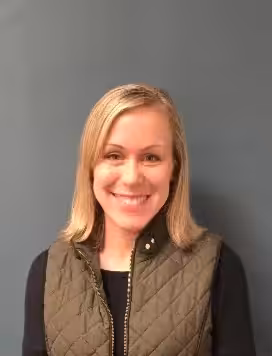 A woman with straight blonde hair, wearing a dark quilted vest over a black top, smiles at the camera while standing in front of a plain blue-gray wall.