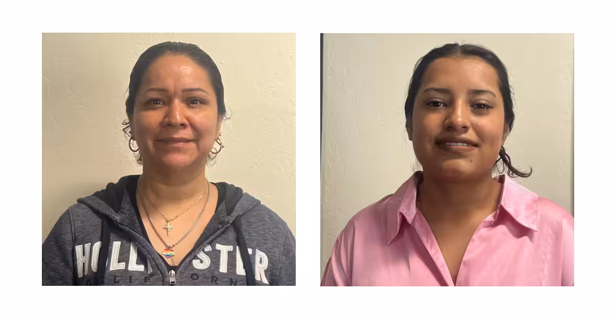 Two women standing against a light-colored wall. The woman on the left wears a dark hoodie and layered necklaces; the woman on the right wears a pink collared shirt and earrings. Both are facing the camera and smiling.