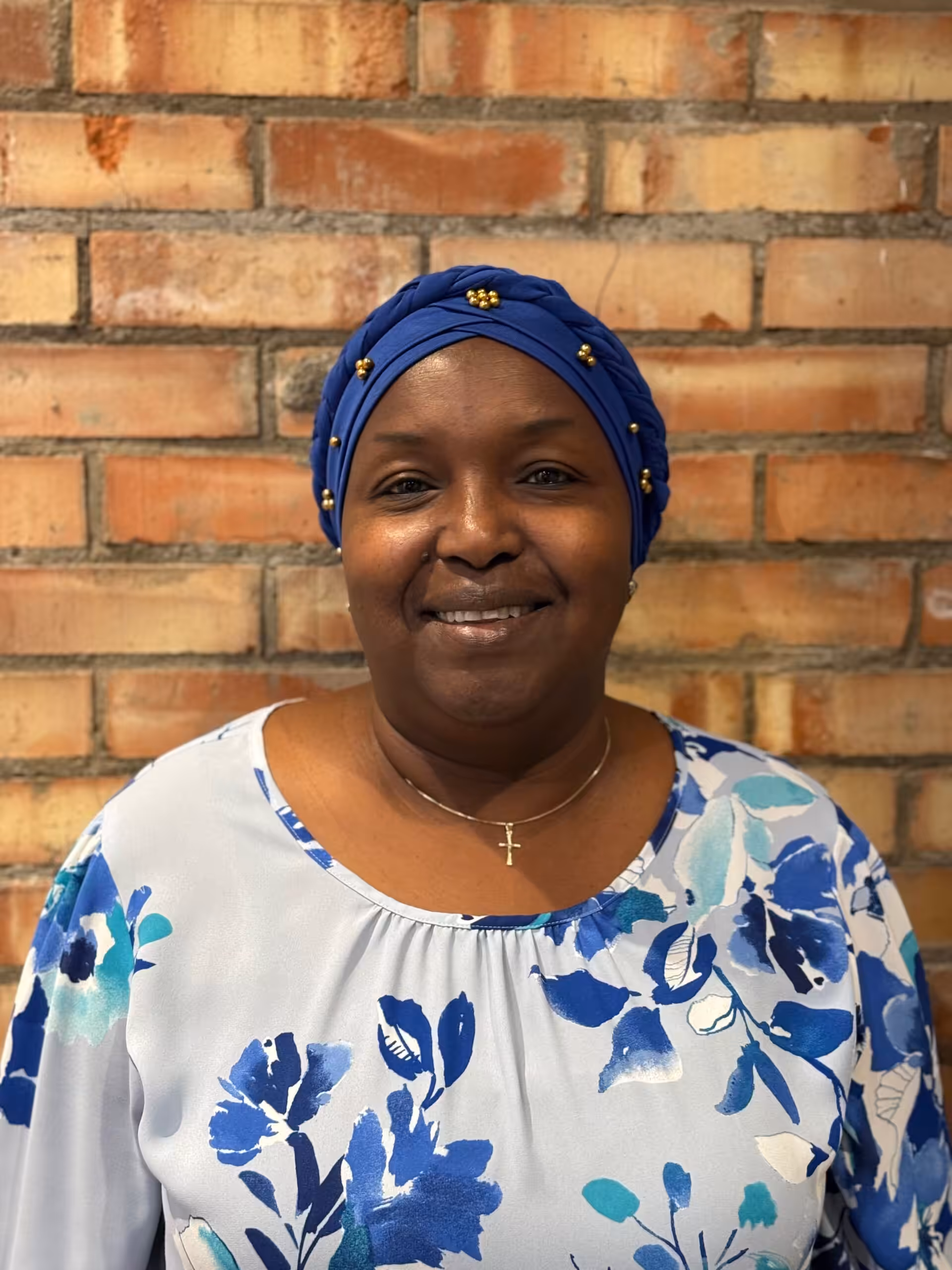 A woman smiles in front of a brick wall. She wears a blue headscarf decorated with small gold embellishments and a light blue floral top. She has a necklace with a cross pendant.