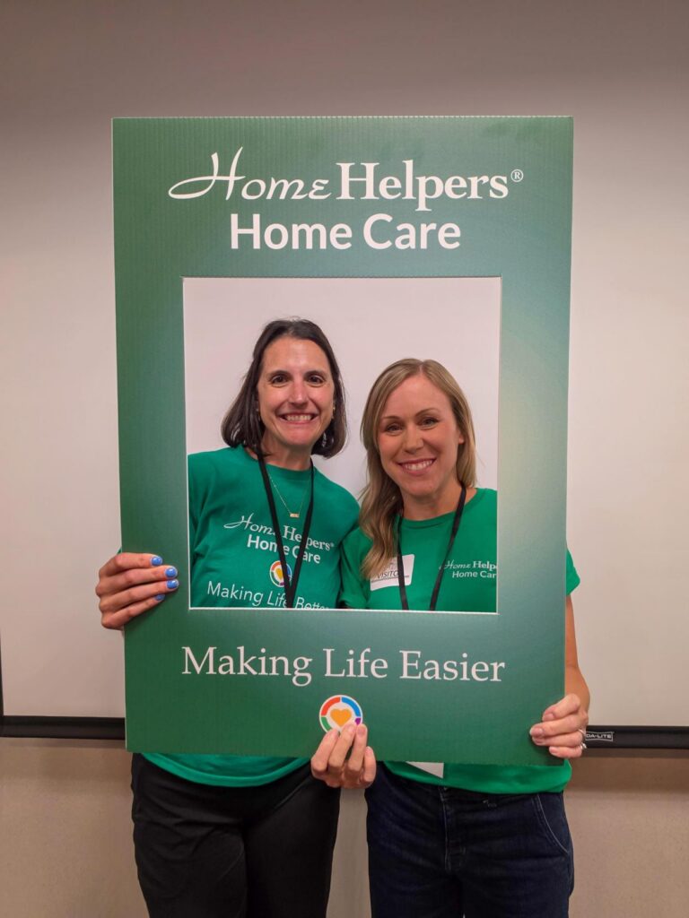 Two women smiling and posing together inside a large Home Helpers Home Care photo frame, both wearing green shirts with the company logo and slogan Making Life Easier printed on them.