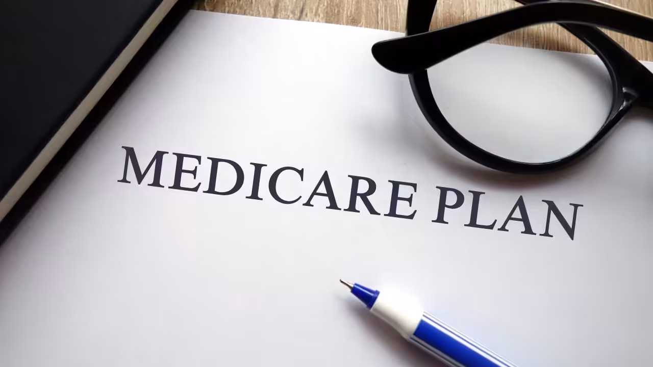 A close-up of a paper titled MEDICARE PLAN next to a pair of black eyeglasses and a blue pen, with part of a black notebook visible on a wooden surface.