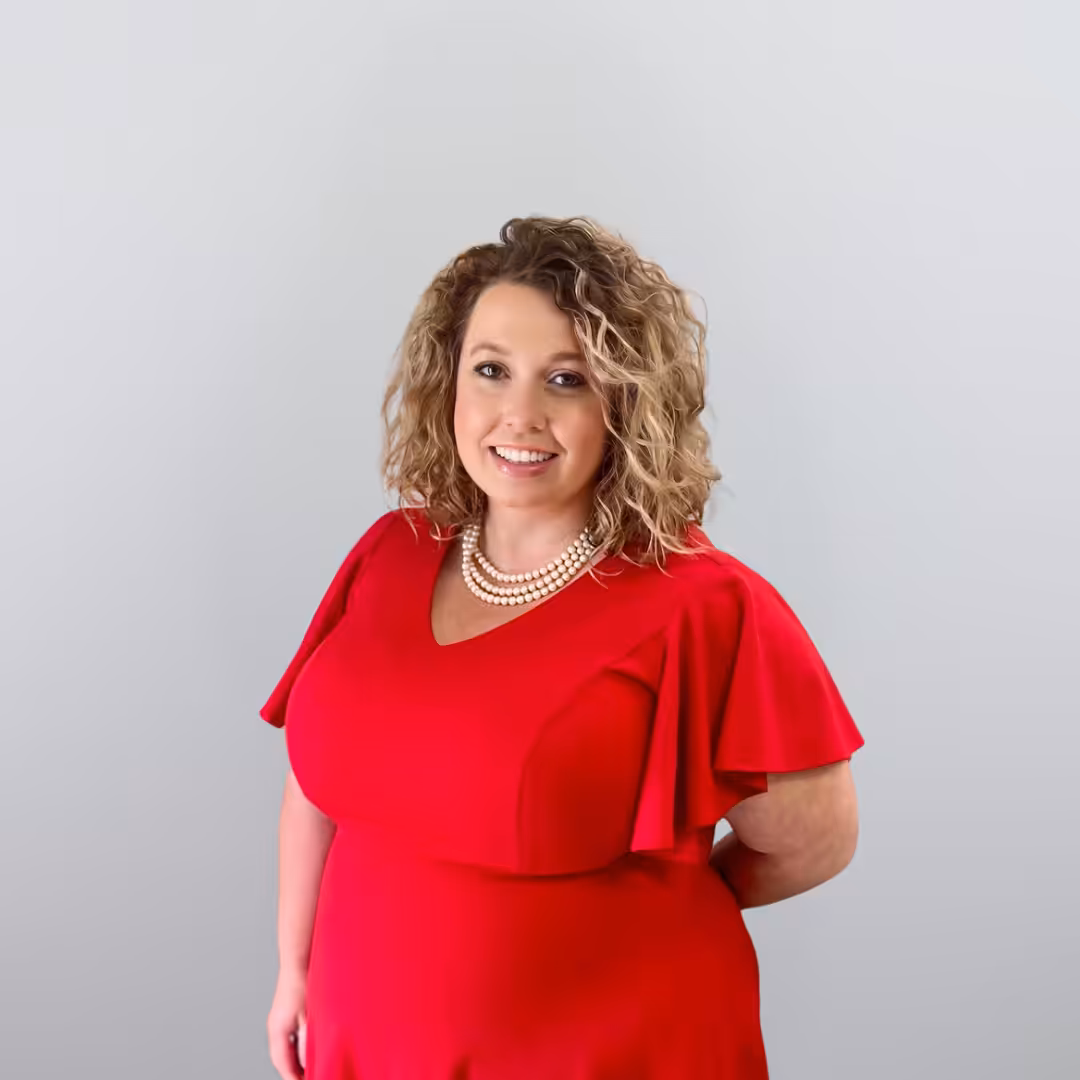 A woman with curly blonde hair wearing a bright red dress and a pearl necklace smiles at the camera against a plain light gray background.