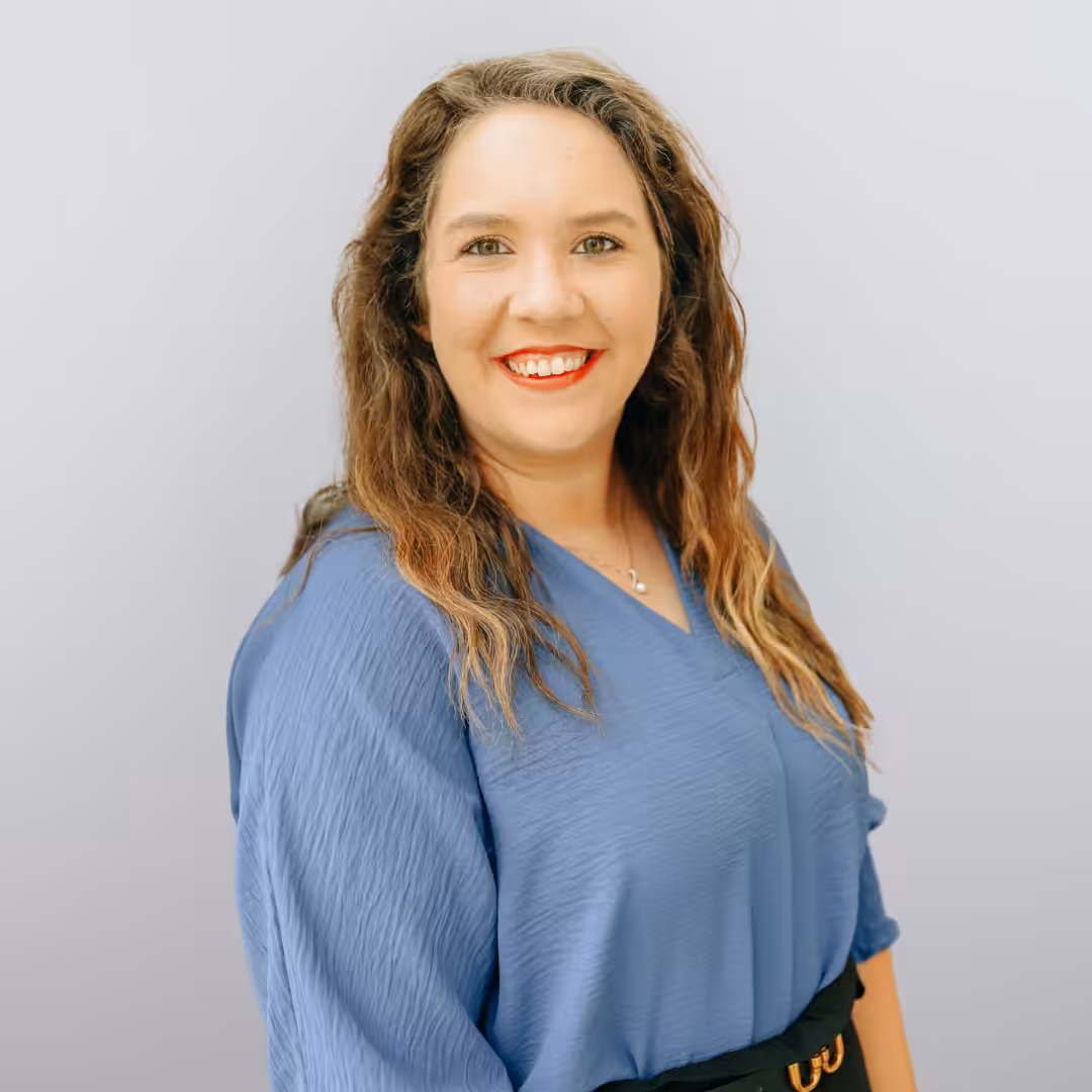 A woman with wavy brown hair wearing a blue blouse and black skirt stands smiling confidently against a light gray background.