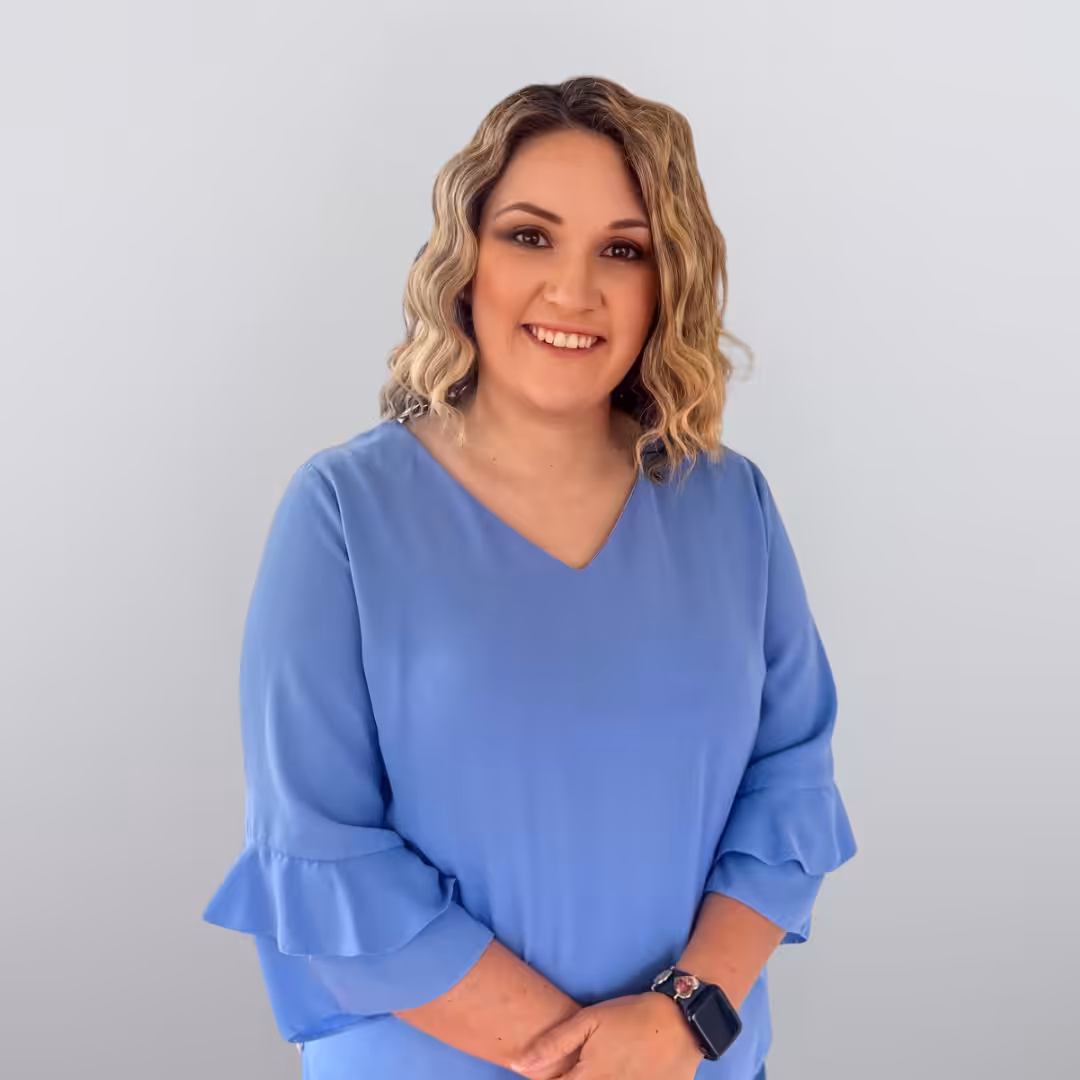 A woman with wavy blonde hair wearing a light blue blouse stands smiling with her hands clasped in front of her against a plain light gray background.